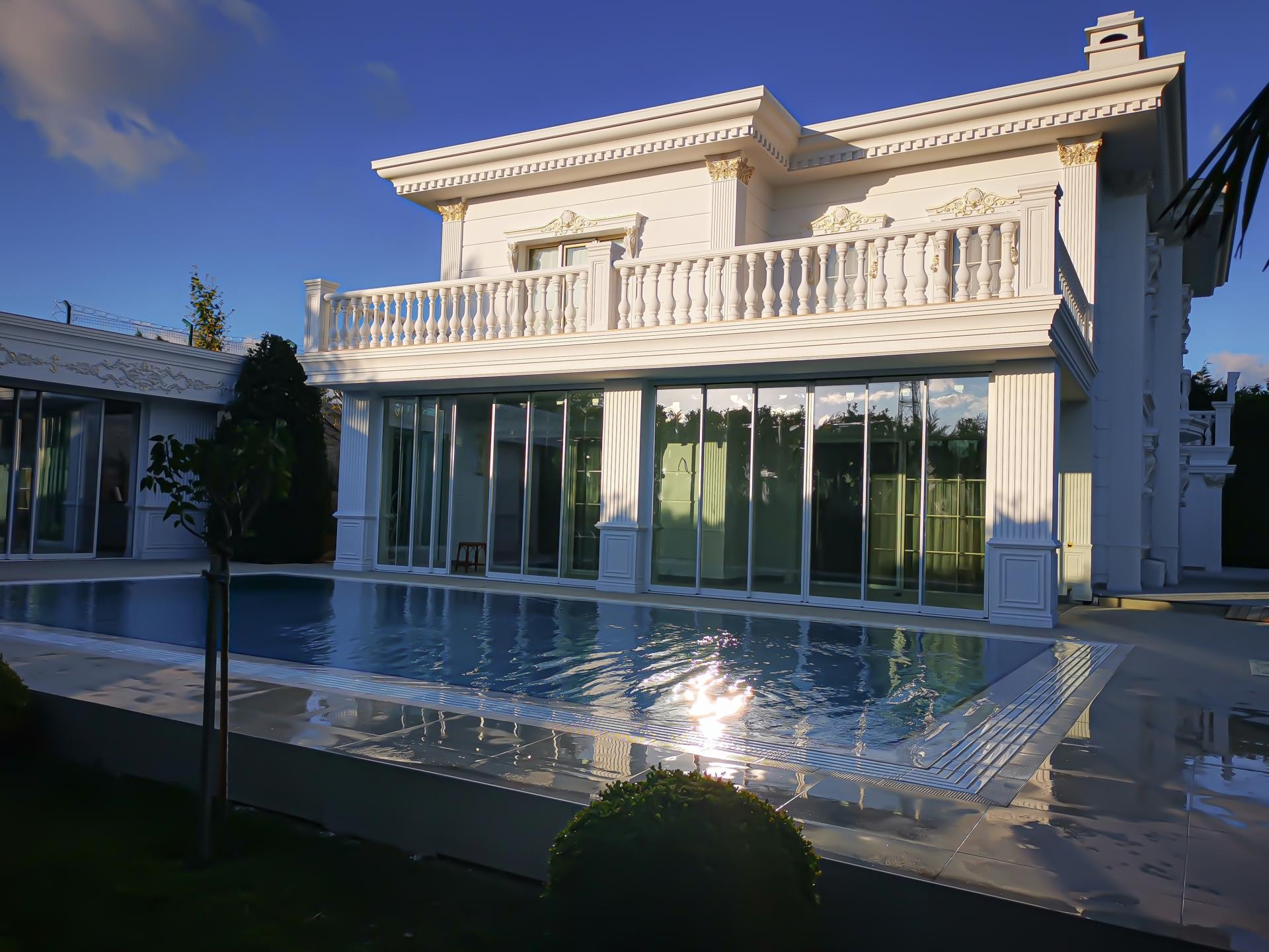 Fațadă albă de vilă clasică cu pilastri din poliuretan, balustrade la etaj și piscină exterioară iluminată.