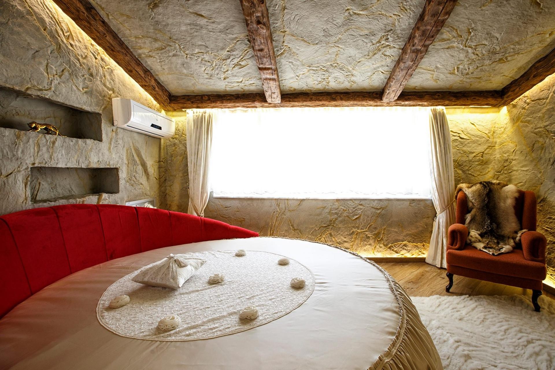Dormitor rustic cu pat rotund roșu, perete din piatră texturată și grinzi decorative din poliuretan P1981C pe tavan.