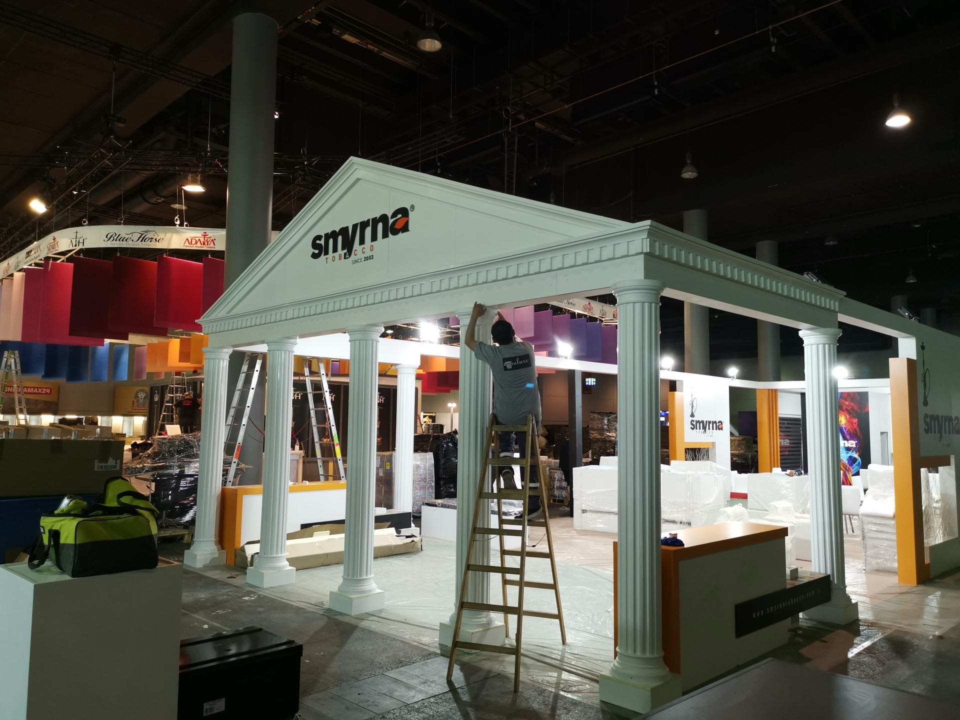 Stand expozițional alb cu coloane clasice din poliuretan și antablament decorativ montat în interiorul unei hale.