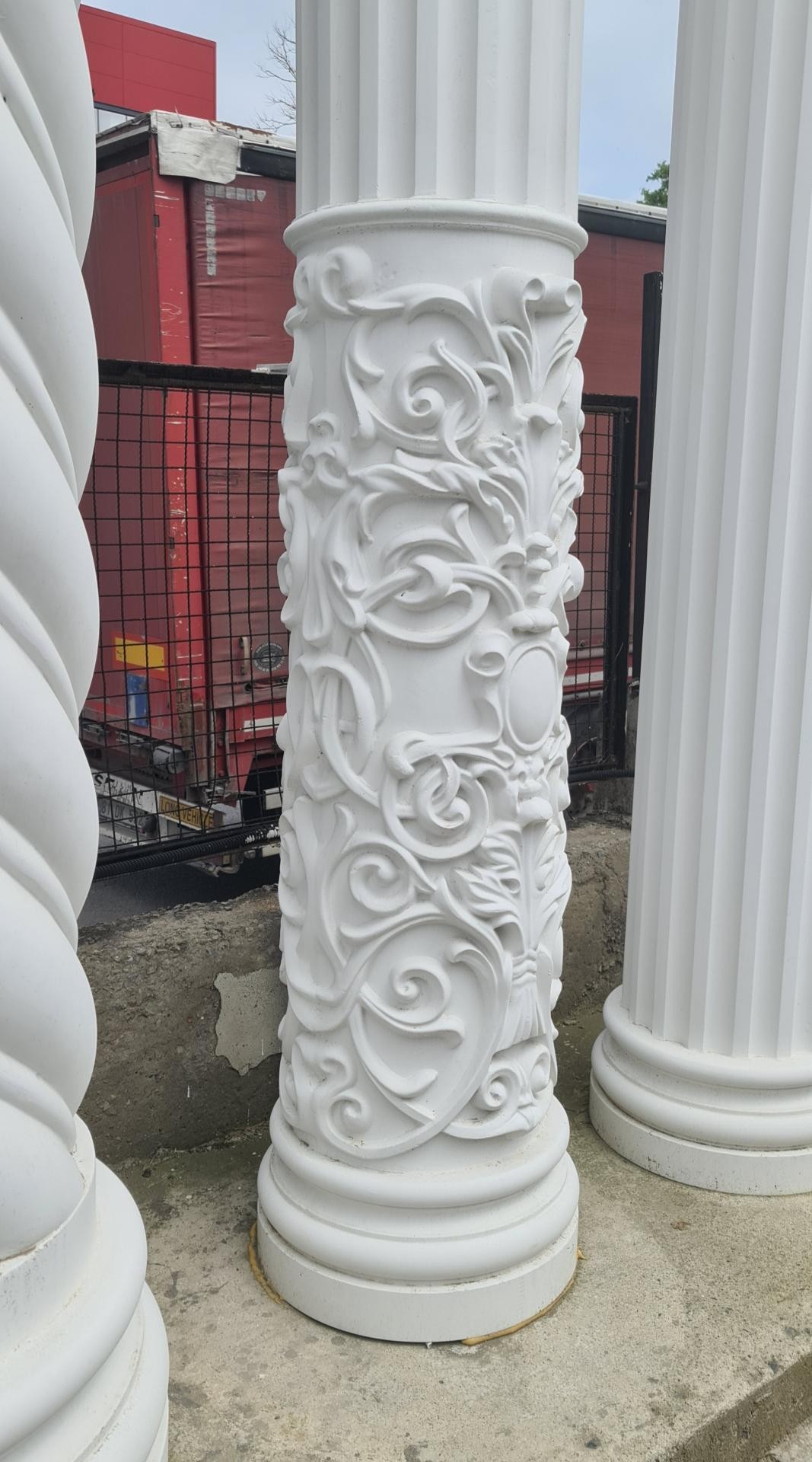 Coloană din poliuretan alb P5540 cu sculptură ornamentală bogată, amplasată lângă coloane canelate pe un suport din beton.