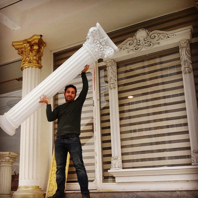 Bărbat susținând o coloană din poliuretan ușoară cu capitul corintic lângă ancadramente decorative de fereastră.
