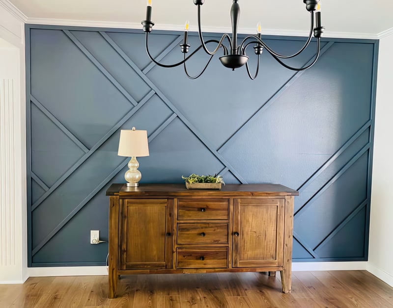 Perete albastru decorat cu profile geometrice din poliuretan P851350, comoda din lemn și candelabru negru.