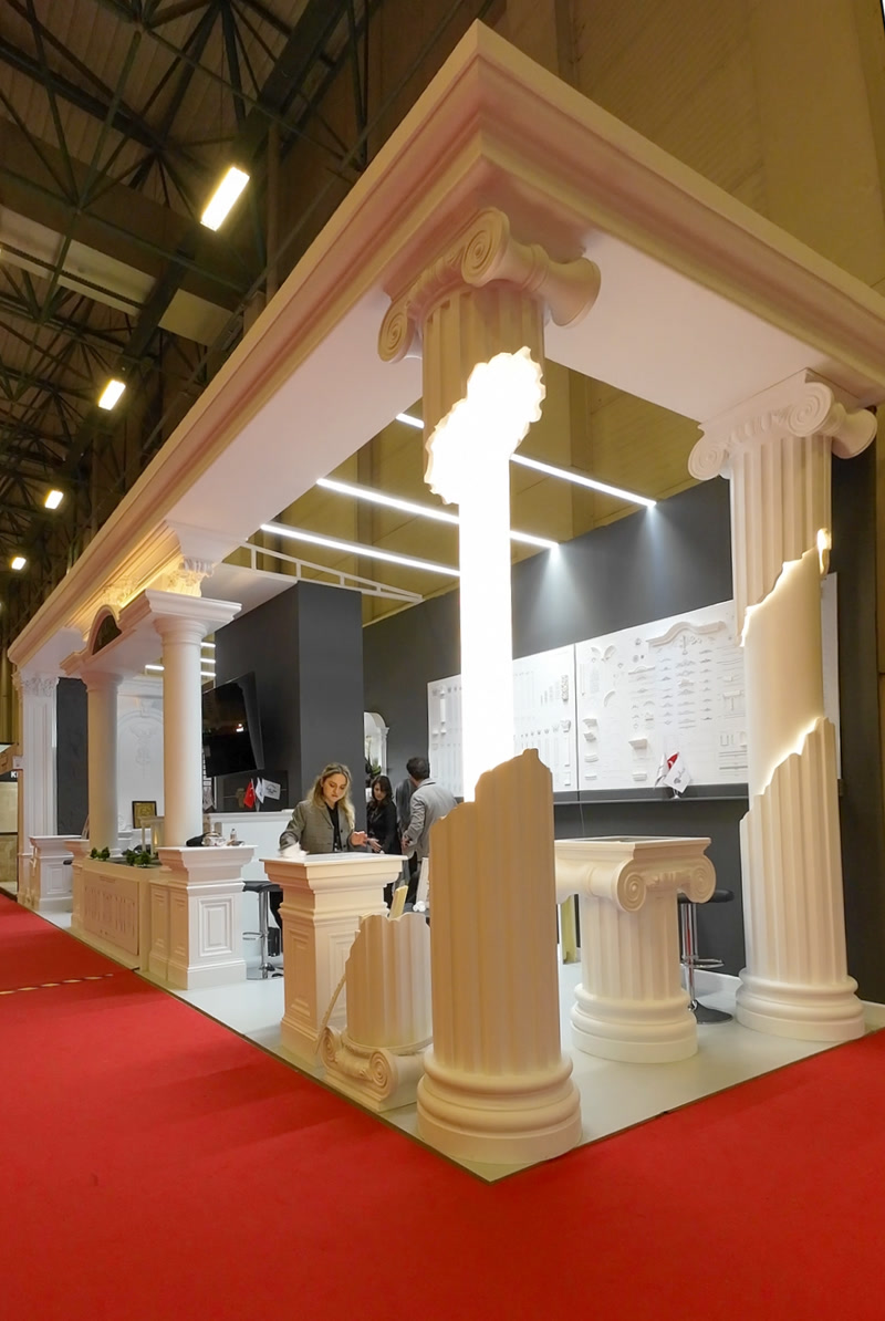 Stand expozițional cu coloane din poliuretan albe tăiate stil antic, iluminat LED integrat și fundal antracit.