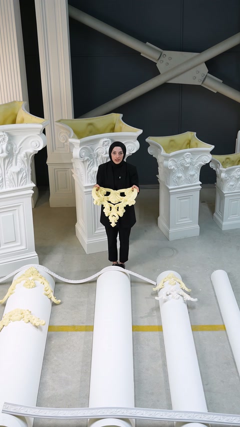 Prezentatoarea arată un ornament galben flexibil între coloane decorative expuse.