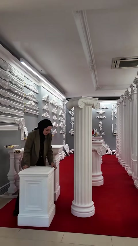 Prezentatoarea examinează un soclu decorativ între coloanele expuse.